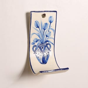 May include: A curved, off-white ceramic wall hook featuring a hand-painted blue design of a vase filled with tulips. The piece has a rectangular shape with a hole at the top for mounting and a blue border outlining the entire edge of the ceramic.