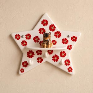May include: A star-shaped wall shelf made of cream-coloured ceramic with a repeating pattern of small red flowers. The shelf features a small, hand-painted ceramic dog figurine sitting on the ledge and three white hooks for hanging items.