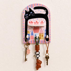 May include: A ceramic wall-mounted key holder featuring a painted black cat arching over a small shelf. The background displays a pastel-coloured town scene with houses. Three white hooks extend from the bottom, each holding a set of keys.