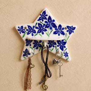 May include: A star-shaped ceramic wall hook rack featuring a white base with hand-painted blue iris flowers and green stems. The piece includes a small horizontal shelf and three white hooks, currently holding a brown lace ribbon and two keychains.