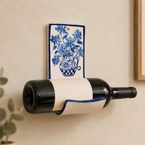 May include: A wall-mounted ceramic wine bottle holder featuring a rectangular backplate decorated with a blue and white floral pitcher illustration. The holder has a curved, off-white cradle that supports a dark glass wine bottle horizontally.