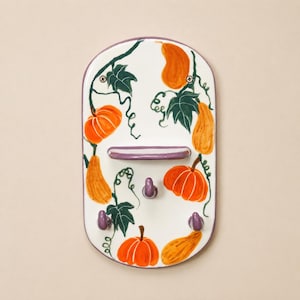 Puede incluir: Un perchero de pared ovalado de cerámica con fondo blanco, decorado con calabazas naranjas, calabacines amarillos y hojas verde oscuro pintadas a mano. Cuenta con un pequeño estante horizontal y tres ganchos morados, con dos tornillos.