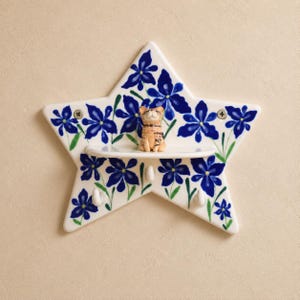 May include: A star-shaped ceramic wall shelf featuring a hand-painted blue floral pattern with green stems. The shelf includes a small horizontal ledge, three white hooks for hanging items, and a small tan cat figurine sitting on the center shelf.