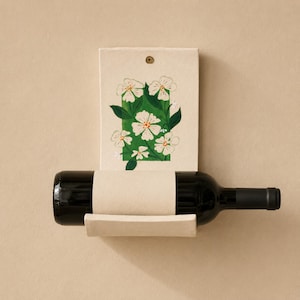 May include: A wall-mounted wine rack made of off-white ceramic with a rectangular backplate featuring a hand-painted green rectangle and white floral design. A dark glass wine bottle rests horizontally in the curved ceramic cradle.