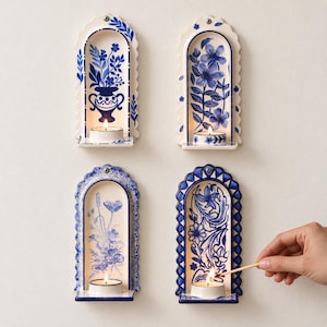 Puede incluir: Cuatro apliques de pared de cerámica en forma de arco con motivos florales azules y blancos. Cada pieza tiene un pequeño estante que sostiene una vela de té encendida. Los diseños incluyen un jarrón, flores y adornos de volutas.