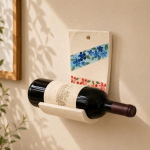 May include: A wall-mounted ceramic wine bottle holder in an off-white finish. The vertical back panel features two diagonal painted bands, one with blue flowers and green leaves, and the other with red flowers. A single screw secures it to the wall.