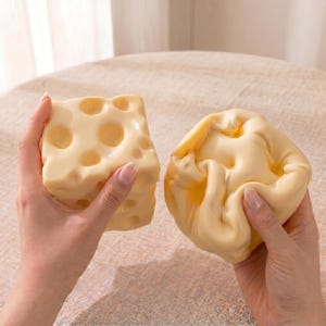Peut inclure: Deux blocs de mousse souples et spongieux de couleur jaune pâle, en forme de fromage suisse. Un bloc conserve une forme cubique avec des indentations circulaires, tandis que l'autre est comprimé et déformé, révélant une texture lisse et souple.