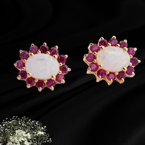 May include: A pair of gold stud earrings, each featuring a central oval-shaped white opal surrounded by a halo of small, round, deep pink rubies. The stones are set in a prong setting, creating a floral-like cluster design on each earring.
