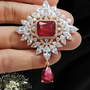 May include: A gold-tone brooch featuring a large rectangular red center stone surrounded by multiple marquise-cut clear crystals. The piece includes a dangling pear-shaped red stone at the bottom, with intricate metalwork framing the stones.