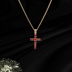 May include: A gold-toned necklace featuring a cross-shaped pendant. The pendant is encrusted with small, round, deep red stones arranged in a vertical and horizontal pattern. The chain is a thin, delicate gold-toned metal link style.
