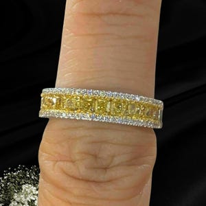May include: A band ring featuring a central row of rectangular yellow stones flanked by two outer rows of small, clear, sparkling stones. The ring has a metallic finish and is designed with a multi-row channel setting for a uniform appearance.