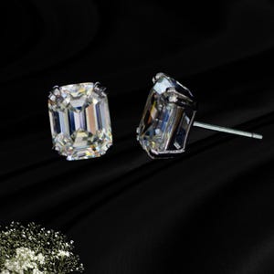 May include: A pair of stud earrings featuring clear, emerald-cut stones set in silver-toned metal prongs. The rectangular stones have faceted surfaces that reflect light, and each earring includes a thin metal post for attachment to the earlobe.
