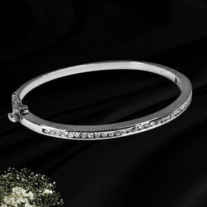 May include: A silver-toned oval hinged bangle bracelet featuring a single row of small, clear, round-cut stones embedded along the front half of the band. The bracelet includes a secure metal clasp mechanism on the left side for opening and closing.