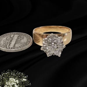 May include: A gold ring featuring a cluster of round, clear stones arranged in a floral pattern. The ring is positioned next to a silver United States dime coin for scale, highlighting the size of the central stone setting on the band.