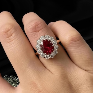 May include: A ring featuring a large, oval-cut deep red gemstone at the center, surrounded by a halo of twelve round, clear, faceted stones. The setting is a polished gold-toned metal band that supports the cluster of stones in a floral arrangement.