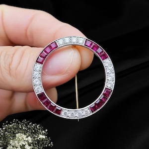 Könnte beinhalten: Eine kreisförmige Brosche mit abwechselnden Abschnitten aus quadratisch geschliffenen, tiefroten Rubinen und rund geschliffenen, klaren Diamanten in einer silberfarbenen Metallfassung. Eine dünne Metallnadel verläuft vertikal durch die Mitte.