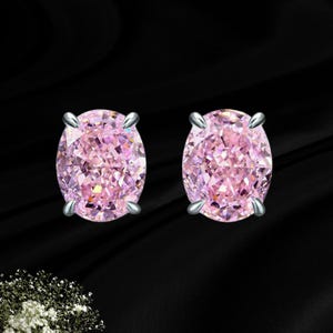 May include: A pair of oval-cut pink gemstone stud earrings, each featuring a faceted stone secured by four silver-toned metal prongs. The stones exhibit a vibrant pink hue with internal light reflections, set against a dark, smooth background.