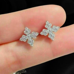 May include: A pair of stud earrings featuring a four-pointed star or flower shape. Each earring is crafted from a silver-toned metal and is encrusted with numerous small, clear, sparkling stones that cover the entire surface of the design.