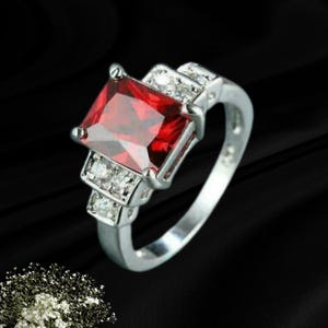 May include: A silver-toned ring featuring a large, rectangular, faceted red gemstone as the centrepiece. The band is accented with two smaller, clear, round-cut stones on each side of the main setting, creating a symmetrical design.