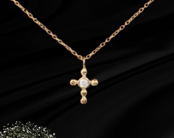 Könnte beinhalten: Eine goldfarbene Halskette mit einer zarten Kette und einem kleinen Kreuzanhänger. Das Kreuz hat in der Mitte einen runden, klaren Stein und vier kleine, abgerundete goldene Akzente an den Enden jedes Arms, was ein schlichtes Schmuckstück ergibt.