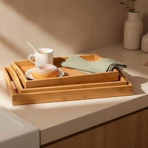 May include: A set of three rectangular bamboo serving trays stacked on top of each other. The top tray holds a white ceramic mug with a small spoon, a white plate with a round donut, and a folded sage green cloth napkin with a textured finish.