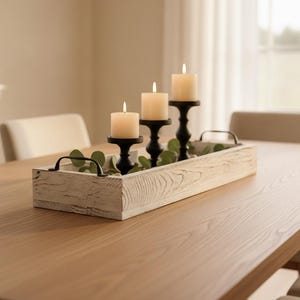 May include: A rectangular white distressed wooden tray with black metal handles containing three black candle holders of varying heights, each topped with a lit cream-colored pillar candle and accented with green eucalyptus leaves.