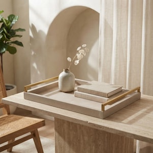 May include: A rectangular beige tray with gold-toned metal handles. The tray holds a small, off-white ceramic vase containing a delicate dried plant stem with translucent, coin-shaped leaves, and two stacked books with light-colored covers.