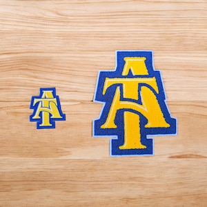 May include: Two embroidered patches featuring the letters A and T intertwined in a collegiate style. The letters are bright yellow with a thick blue border. The patches are shown in two different sizes, both displaying the same design.