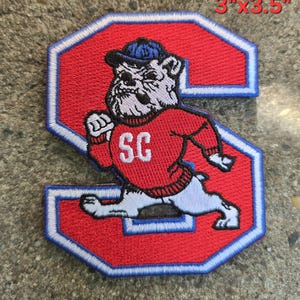 Pode incluir: Um emblema bordado em vermelho e azul com o formato da letra S, apresentando um bulldog usando um boné azul e um suéter vermelho com as letras SC. O emblema mede 7,62 cm por 8,89 cm e tem uma borda branca.