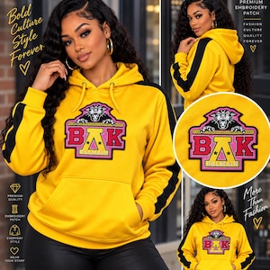May include: A bright yellow hooded sweatshirt featuring a large embroidered patch on the chest. The patch displays a black panther above the letters BAK in pink and yellow, with the word BALMAIN below. It has black side stripes and a front pocket.