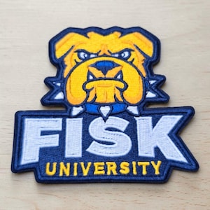 Puede incluir: Un parche bordado que presenta una cabeza de bulldog amarilla con un collar azul y púas blancas, colocado sobre el texto FISK UNIVERSITY en letras blancas y amarillas sobre un fondo azul oscuro con un borde azul oscuro.