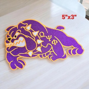May include: A purple embroidered patch in the shape of a bulldog with white teeth and collar spikes. The design features the Greek letters Omega, Psi, and Phi in gold thread. The patch measures 5 inches by 3 inches as indicated by the text.