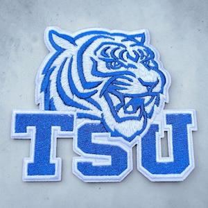 May include: An embroidered patch featuring a white tiger head with blue stripes and a snarling expression, positioned above the bold, block-lettered text TSU in blue with a white border. The entire design is outlined in white thread.