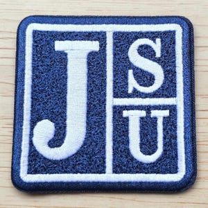 Può includere: Una toppa ricamata quadrata con sfondo blu scuro, bordo bianco e scritte bianche. Il design è diviso in due sezioni, con una grande lettera J sulla sinistra e le lettere S e U impilate sul lato destro della toppa.