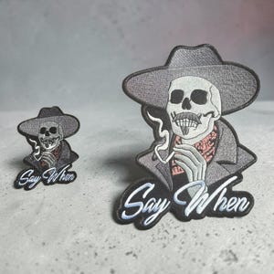 Puede incluir: Dos parches bordados que muestran un esqueleto con sombrero de ala ancha, bigote y perilla. El esqueleto sostiene una pipa humeante, lleva un pañuelo estampado y el texto Say When está bordado en letra cursiva blanca en la parte inferior.