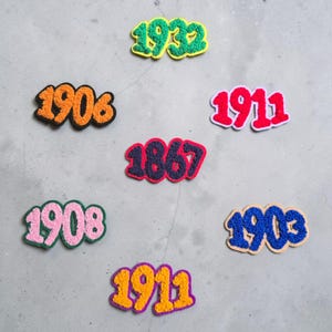 May include: Seven chenille patches featuring the numbers 1932 in green and yellow, 1906 in orange and black, 1911 in red and white, 1867 in dark blue and red, 1908 in pink and green, 1903 in blue and tan, and 1911 in yellow and purple.