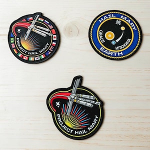 May include: Three circular embroidered patches featuring space-themed designs. Two patches depict a spacecraft with a red trail and the text Project Hail Mary. One patch features a blue border with the text Hail Mary Grace Earth and symbols.