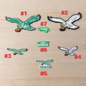 May include: A collection of seven embroidered patches featuring eagle designs and text. Patches include green and white eagles holding footballs, a white eagle, and text patches reading #1, #2, #3, #4, #5, #6, #7, Philly, Go Birds, and Eagles.