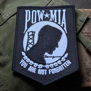 May include: A black and white embroidered patch featuring the text POW MIA at the top and YOU ARE NOT FORGOTTEN at the bottom. The central design shows a white circular background with a black silhouette of a person and a guard tower.