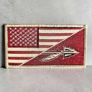 Puede incluir: Un parche rectangular bordado con un diseño dividido. El lado izquierdo muestra una bandera estadounidense roja y blanca con un cantón azul lleno de estrellas blancas. El lado derecho es rojo sólido con un icono de punta de flecha y pluma bordado.