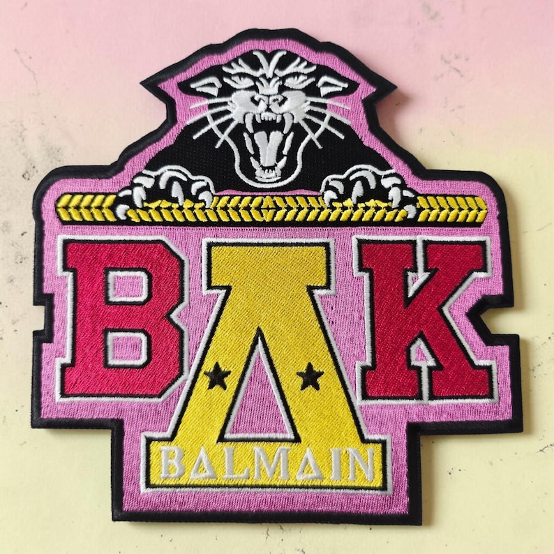 Pode incluir: Um emblema bordado apresentando uma cabe&ccedil;a de pantera preta com detalhes brancos acima de uma borda de corda amarela. Abaixo, as letras B, &Delta; e K s&atilde;o exibidas em rosa e amarelo, com a palavra BALMAIN impressa em branco dentro do s&iacute;mbolo delta amarelo.