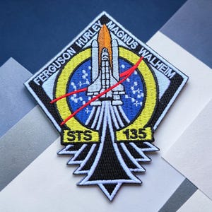 May include: A diamond-shaped embroidered patch featuring the NASA STS-135 mission emblem. It displays a space shuttle launching into a blue circular field with stars, surrounded by a yellow ring. Text reads FERGUSON HURLEY MAGNUS WALHEIM STS 135.