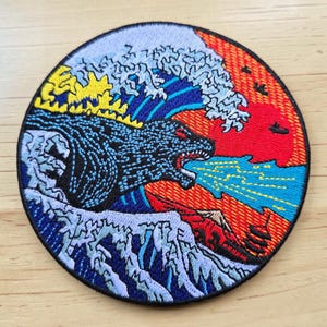 May include: A circular embroidered patch featuring a black monster with yellow dorsal fins breathing blue fire. The design includes a large crashing wave in shades of blue and white, set against a vibrant red and orange sunset background.