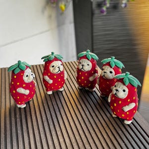 May include: Five small figurines shaped like dogs wearing red strawberry costumes with green leaves on top. Each figure has a cream-coloured face, small ears, and a red body covered in yellow dots, standing upright on a dark brown slatted surface.