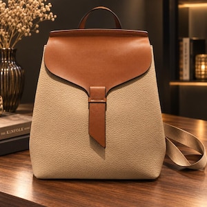 May include: A structured backpack featuring a beige pebbled texture body and a smooth tan leather flap closure. The bag includes a top handle, a vertical leather strap detail on the front, and matching beige fabric shoulder straps for carrying.