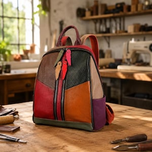 May include: A multi-colored leather backpack featuring a patchwork design in shades of red, orange, brown, black, purple, and olive green. The bag includes a top handle, two shoulder straps, and a front vertical zippered pocket with colorful tags.