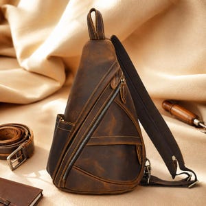 May include: A brown leather sling backpack featuring a triangular shape, a single adjustable shoulder strap, multiple zippered compartments with silver-toned hardware, and visible stitching details along the edges of the leather panels.