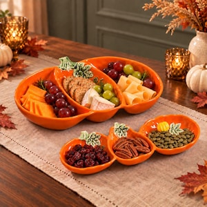 Puede incluir: Dos fuentes de cerámica naranja con forma de calabaza y detalles de hojas verdes. La fuente grande tiene tres secciones con queso, galletas, uvas y brie. La fuente pequeña tiene tres secciones con arándanos secos, nueces pecanas y semillas de calabaza.