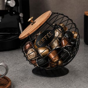 Könnte beinhalten: Ein kugelförmiger Kaffeekapselhalter aus schwarzem Draht mit einem Holzdeckel und einem zentralen schwarzen ovalen Schild, das das Wort COFFEE in goldener Schrift trägt. Der Halter ist mit verschiedenen metallischen Kapseln gefüllt und steht auf einem Fuß.