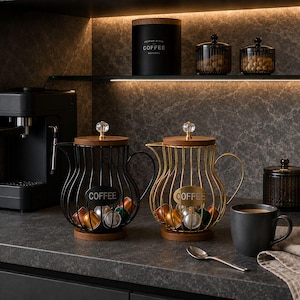 Può includere: Due porta capsule da caffè in filo metallico con basi e coperchi in legno, uno nero e uno dorato, ciascuno con una targhetta ovale centrale con la scritta COFFEE. I supporti contengono capsule assortite e pomelli in cristallo.